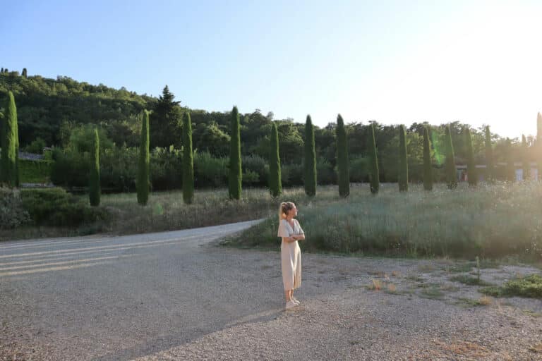 A woman in a light summer dress stands on a gravel path in front of a long row of slender cypress trees in the green garden of Abbadia Wine Relais in Tuscany.