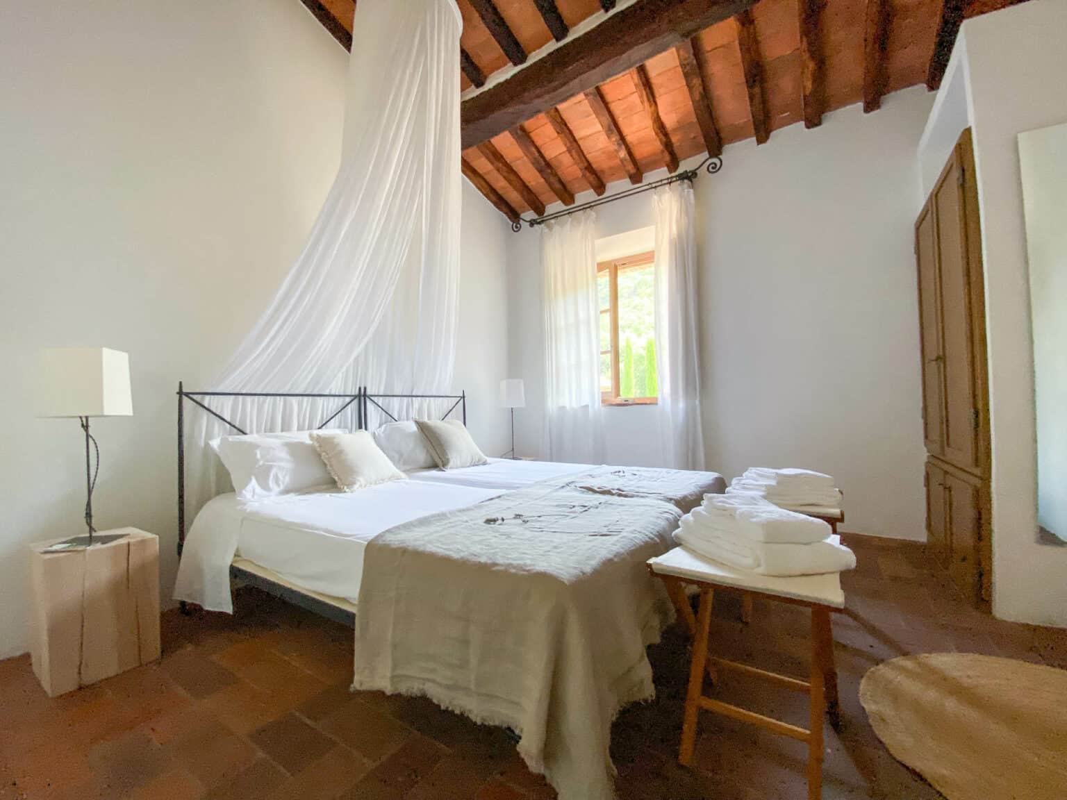 Twin beds with natural linen fabrics, white canopy drapes, and handcrafted terracotta floors in a rustic Tuscan boutique hotel room.